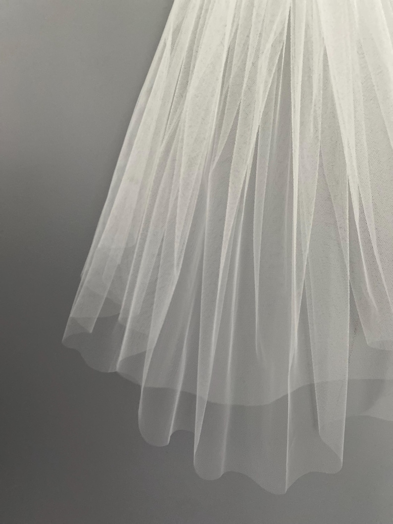May include: A white tulle bridal veil with a single layer of fabric. The veil is pleated and has a scalloped edge.