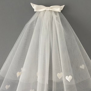 May include: A white bridal veil with a satin bow at the top. The veil is made of sheer fabric and adorned with small, heart-shaped appliqu&eacute;s. The veil is designed to be worn for a wedding.