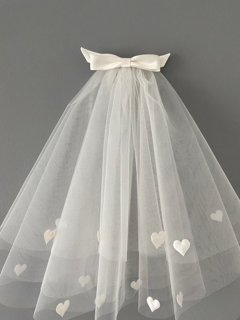 May include: A white bridal veil with a satin bow at the top. The veil is made of sheer fabric and adorned with small, heart-shaped appliqu&eacute;s. The veil is designed to be worn for a wedding.