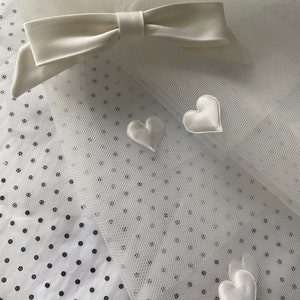 May include: Ivory tulle fabric with a bow and heart accents. The bow is made of a satin-like material and is positioned at the top of the tulle. Three white satin hearts are scattered across the tulle. The background is a white fabric with black polka dots.