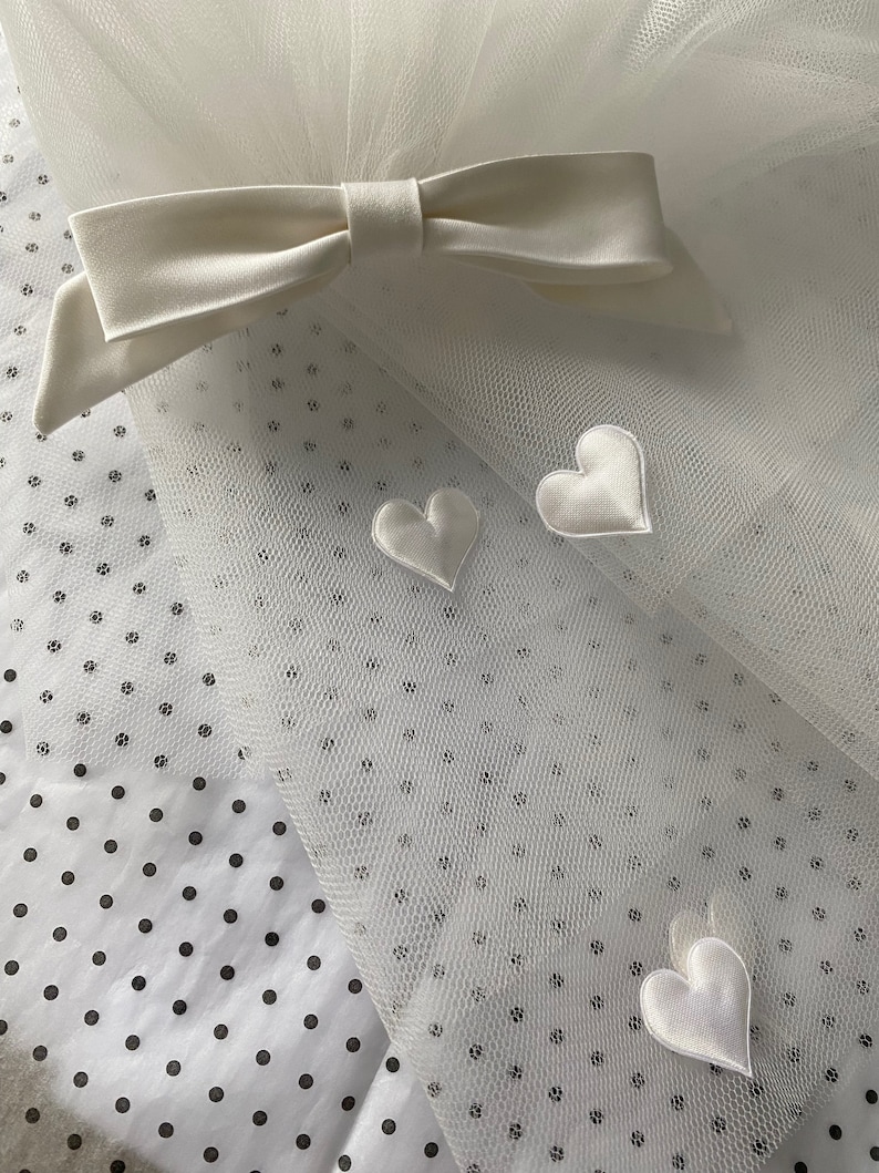 May include: Ivory tulle fabric with a bow and heart accents. The bow is made of a satin-like material and is positioned at the top of the tulle. Three white satin hearts are scattered across the tulle. The background is a white fabric with black polka dots.