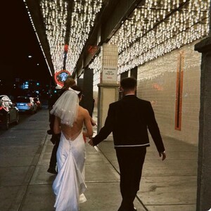 May include: A couple walks down a sidewalk at night, the bride in a white backless wedding dress and veil, the groom in a black suit. Overhead, strings of lights illuminate the scene. The setting appears to be a city street.
