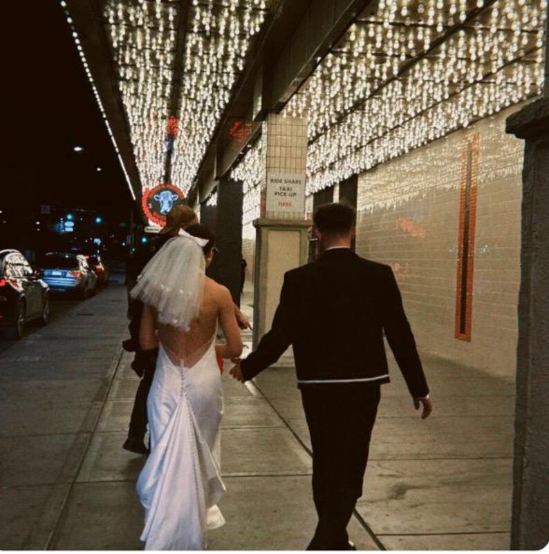 May include: A couple walks down a sidewalk at night, the bride in a white backless wedding dress and veil, the groom in a black suit. Overhead, strings of lights illuminate the scene. The setting appears to be a city street.