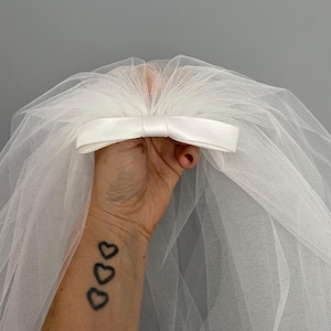 May include: A white tulle bridal veil with a satin bow. The veil is held in a hand with a tattoo of three hearts.