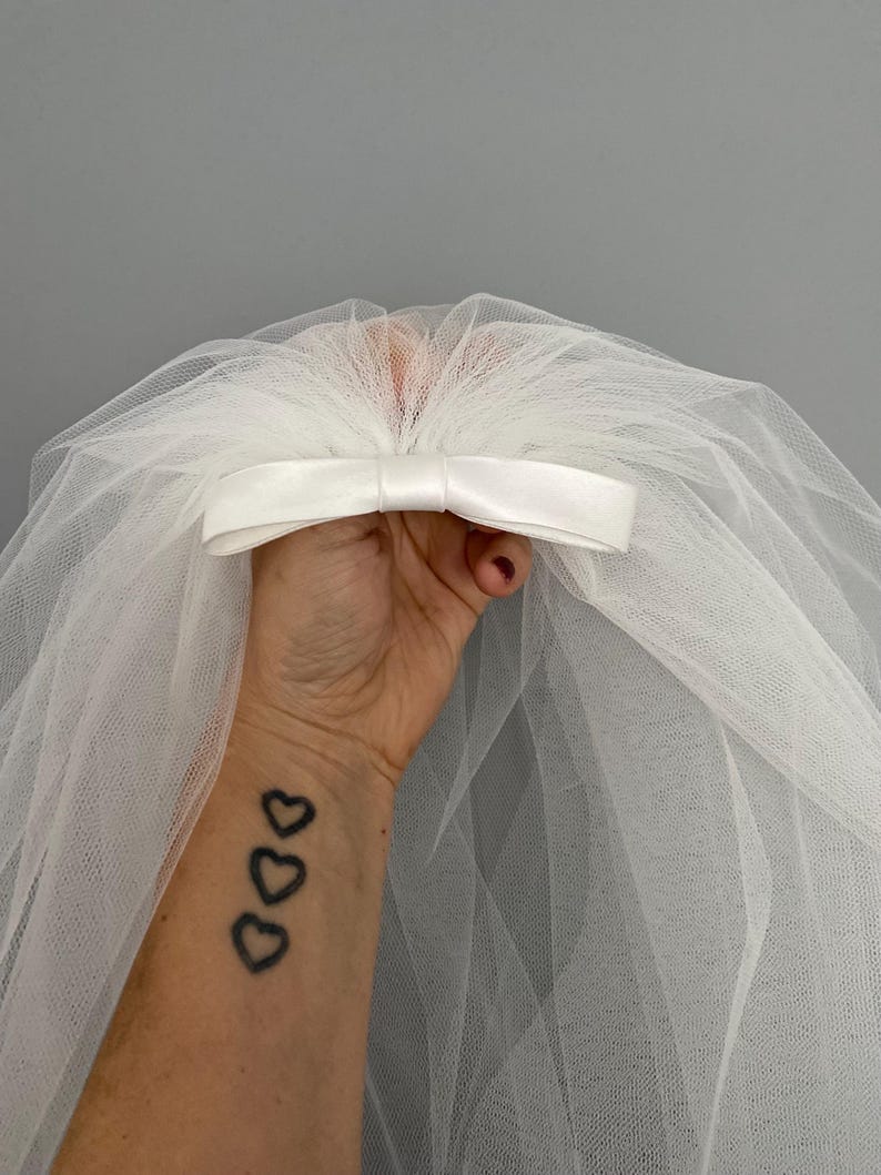 May include: A white tulle bridal veil with a satin bow. The veil is held in a hand with a tattoo of three hearts.