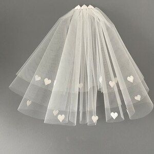 May include: A white tulle bridal veil with multiple layers. The veil is adorned with small, white heart-shaped appliqu&eacute;s. The veil is gathered at the top and cascades down in a layered fashion, creating a soft, romantic look.