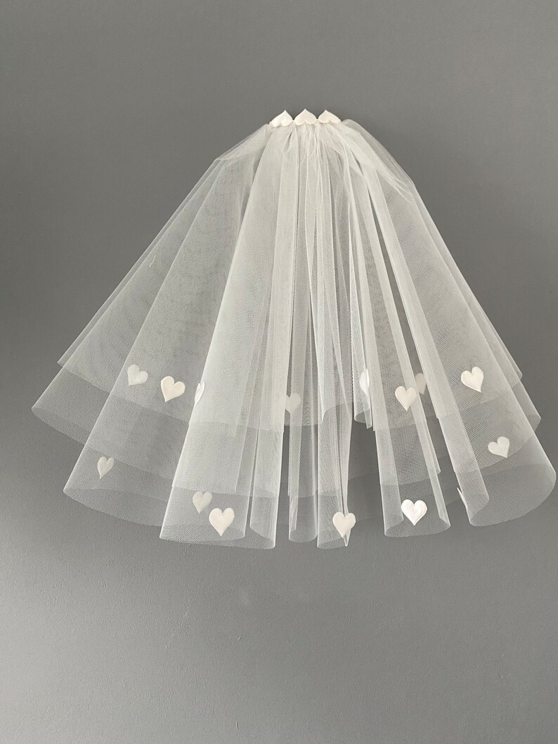 May include: A white tulle bridal veil with multiple layers. The veil is adorned with small, white heart-shaped appliqu&eacute;s. The veil is gathered at the top and cascades down in a layered fashion, creating a soft, romantic look.