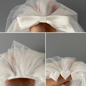 May include: Ivory tulle bridal veil with a satin bow and heart accents. The veil is made of a soft, sheer fabric and features a delicate design. The bow is centered at the top, and the heart accents are along the bottom edge.