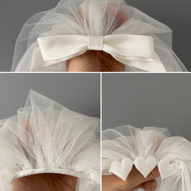 May include: Ivory tulle bridal veil with a satin bow and heart accents. The veil is made of a soft, sheer fabric and features a delicate design. The bow is centered at the top, and the heart accents are along the bottom edge.