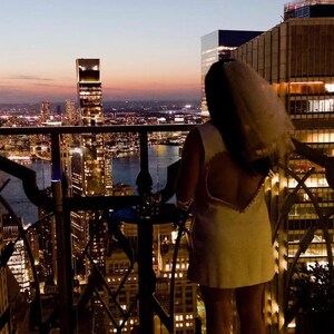 May include: A woman in a white dress with a heart-shaped open back stands on a balcony overlooking a city skyline at dusk. The dress is paired with a veil. The city lights are illuminated in the background.