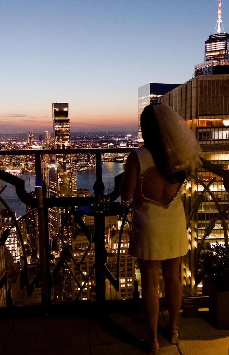 May include: A woman in a white dress with a heart-shaped open back stands on a balcony overlooking a city skyline at dusk. The dress is paired with a veil. The city lights are illuminated in the background.