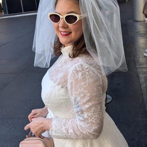 May include: A woman wearing a white lace wedding dress with a white veil and white sunglasses. She is holding a pink handbag with a white dog design.