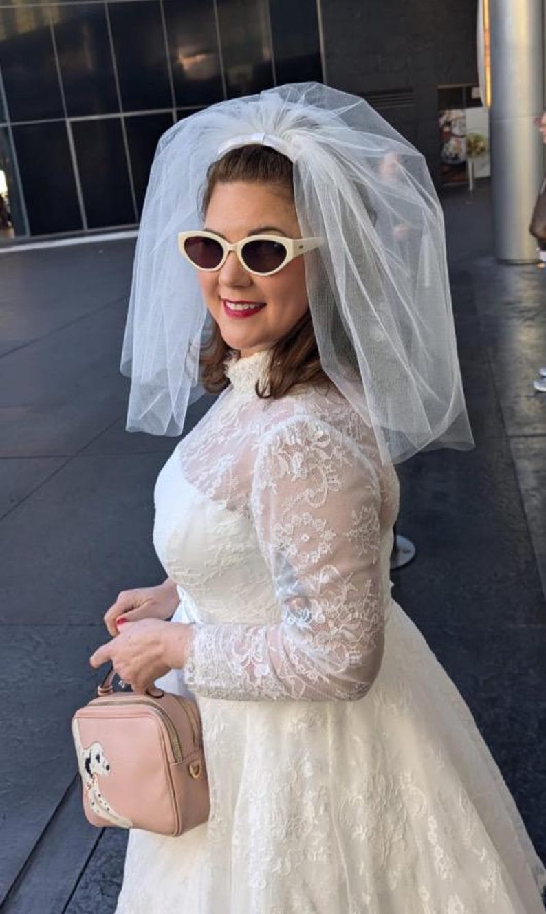 May include: A woman wearing a white lace wedding dress with a white veil and white sunglasses. She is holding a pink handbag with a white dog design.