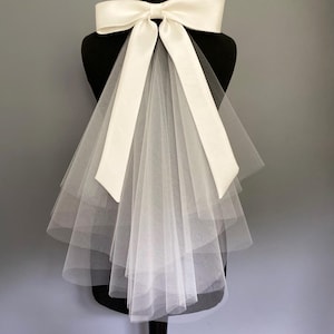 May include: A white bridal veil featuring a large ivory satin bow. Long ribbons flow down over layers of sheer, white tulle. The veil is displayed on a black mannequin against a grey background.
