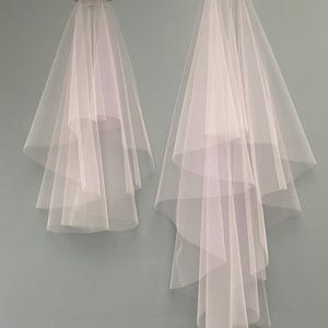 May include: Two white bridal veils. One veil has a pink bow at the top, and the other has a comb. Both veils are made of sheer, layered fabric and hang against a light blue-gray wall.