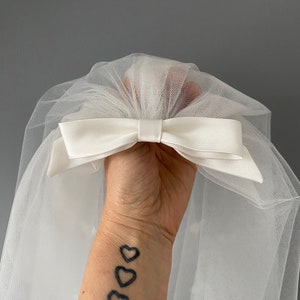 May include: A white tulle veil with a satin bow. The bow is centered and adds a classic touch to the bridal accessory. The veil is held up by a hand.