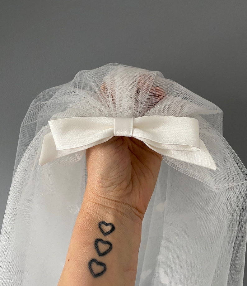 May include: A white tulle veil with a satin bow. The bow is centered and adds a classic touch to the bridal accessory. The veil is held up by a hand.