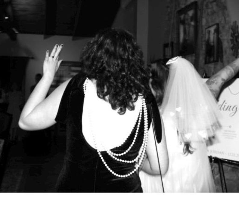 May include: Black velvet dress with a white back panel and a pearl necklace design. A bride in a white wedding dress and veil is visible. The image is in black and white.