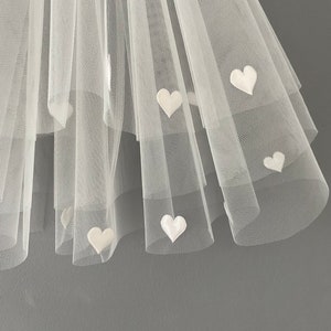 May include: Close-up of a white tulle fabric with multiple layers. Small, white heart-shaped appliqu&eacute;s are scattered across the fabric. The tulle is semi-transparent, and the background is a solid gray.