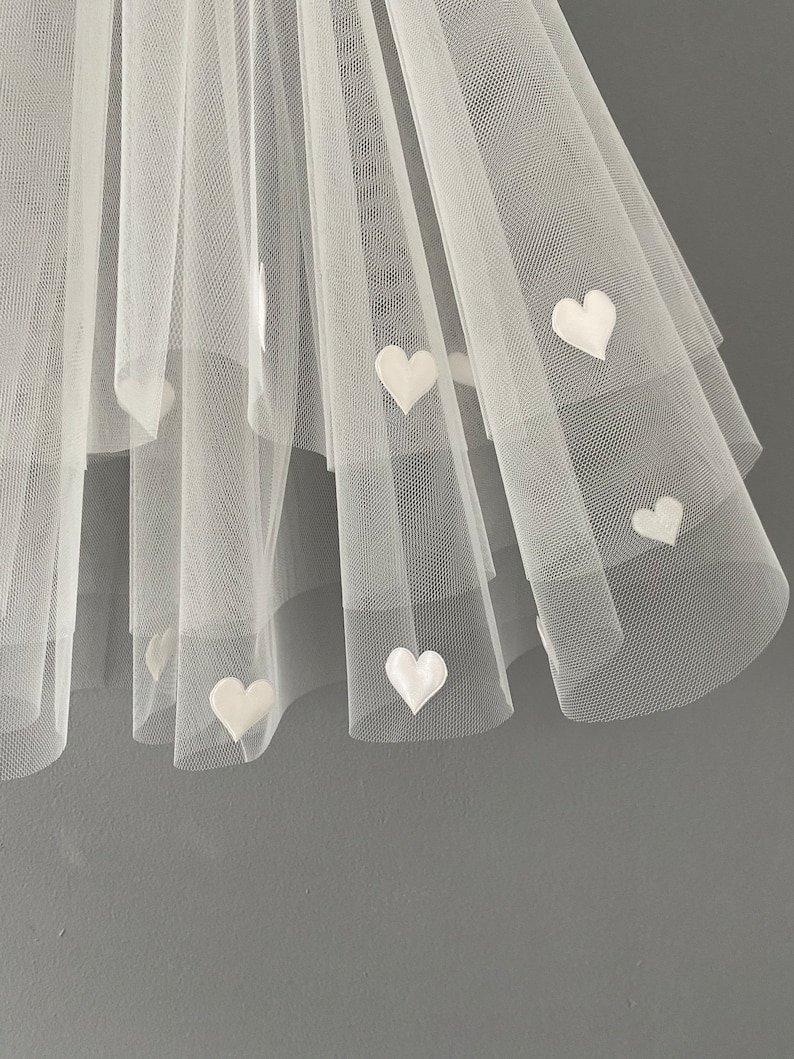 May include: Close-up of a white tulle fabric with multiple layers. Small, white heart-shaped appliqu&eacute;s are scattered across the fabric. The tulle is semi-transparent, and the background is a solid gray.