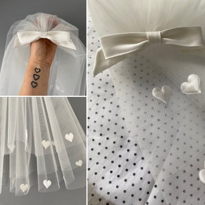 May include: A white tulle veil with satin bow and heart accents. The veil features a large satin bow at the top and small white satin hearts scattered across the tulle. The veil is designed for special occasions.