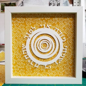May include: A framed paper art piece featuring a white, layered spiral design with Hebrew text, set against a yellow floral patterned background. The artwork is encased in a white square frame, creating a striking contrast.