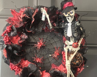 May include: A black and red Halloween wreath with a skeleton wearing a top hat and tie. The wreath is decorated with black roses, red berries, and a spiderweb with spiders.