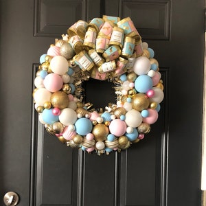 May include: A pink and blue Christmas wreath with gold ornaments and a gold ribbon with a horse design. The wreath is hanging on a black door with the word "Welcome" written on it.