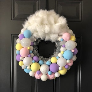 May include: A pastel-colored Christmas wreath with white faux fur, silver tinsel, and a variety of pastel-colored ornaments. The wreath is hanging on a black door.