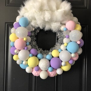 May include: A pastel-colored Christmas wreath with white faux fur, silver tinsel, and a variety of round ornaments in shades of pink, yellow, blue, and purple.