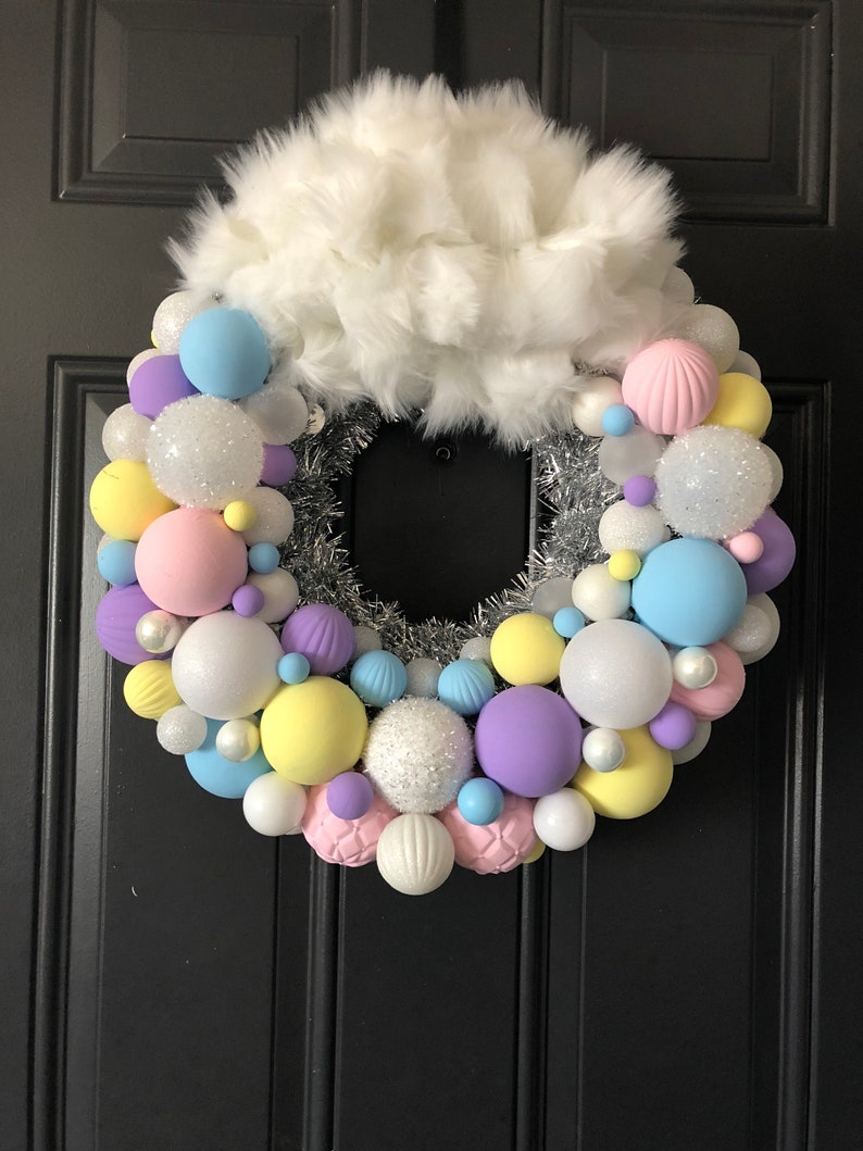 May include: A pastel-colored Christmas wreath with white faux fur, silver tinsel, and a variety of round ornaments in shades of pink, yellow, blue, and purple.
