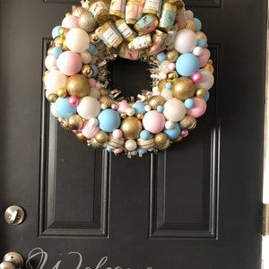 May include: A gold, pink, and blue Christmas wreath with a large bow and the words "Welcome Home" on the ribbon. The wreath is decorated with ornaments and tinsel.