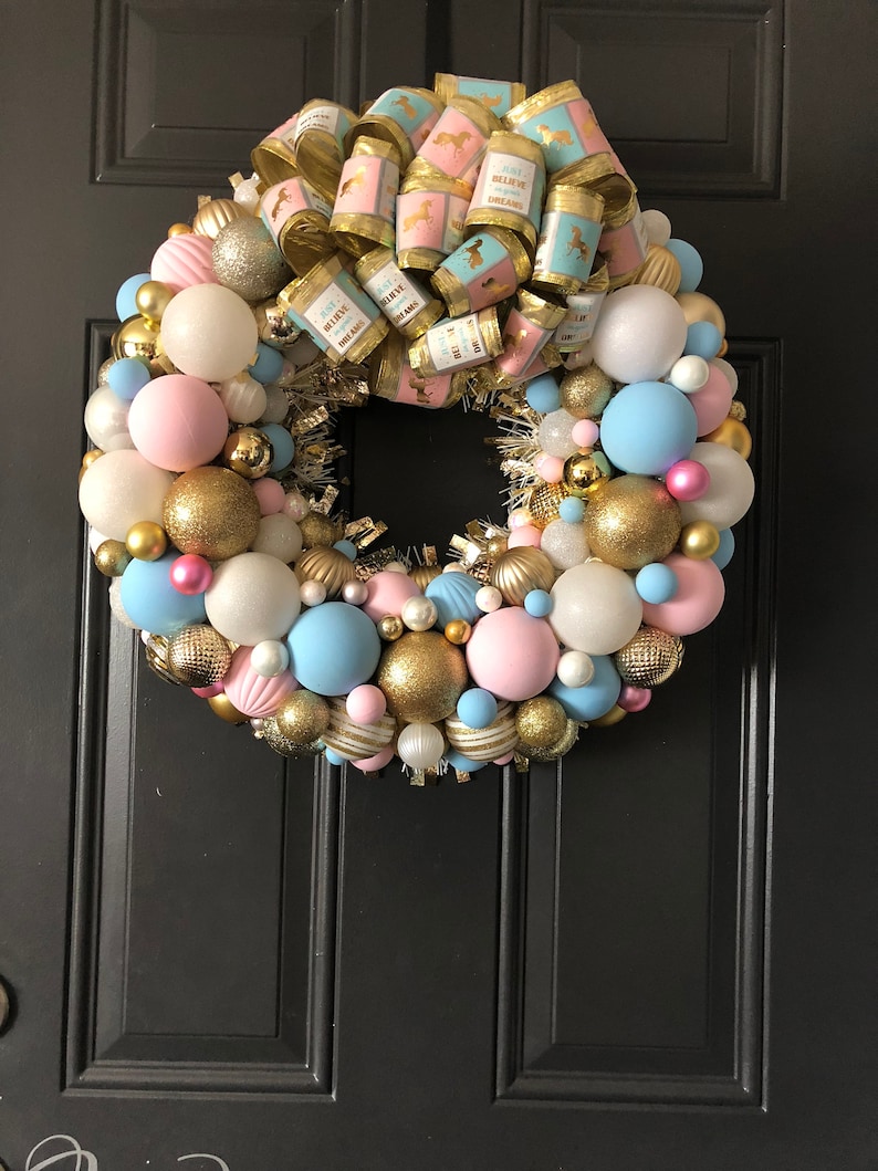 May include: A pink, blue, and gold Christmas wreath with a bow that says "Just Believe in Your Dreams".