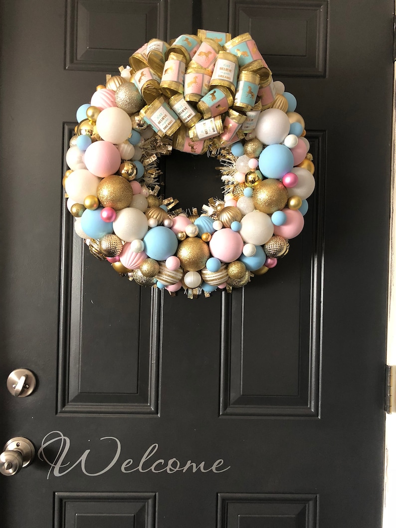 May include: A pink and blue Christmas wreath with gold ornaments and a gold ribbon with a horse design. The wreath is hanging on a black door with the word "Welcome" written on it.