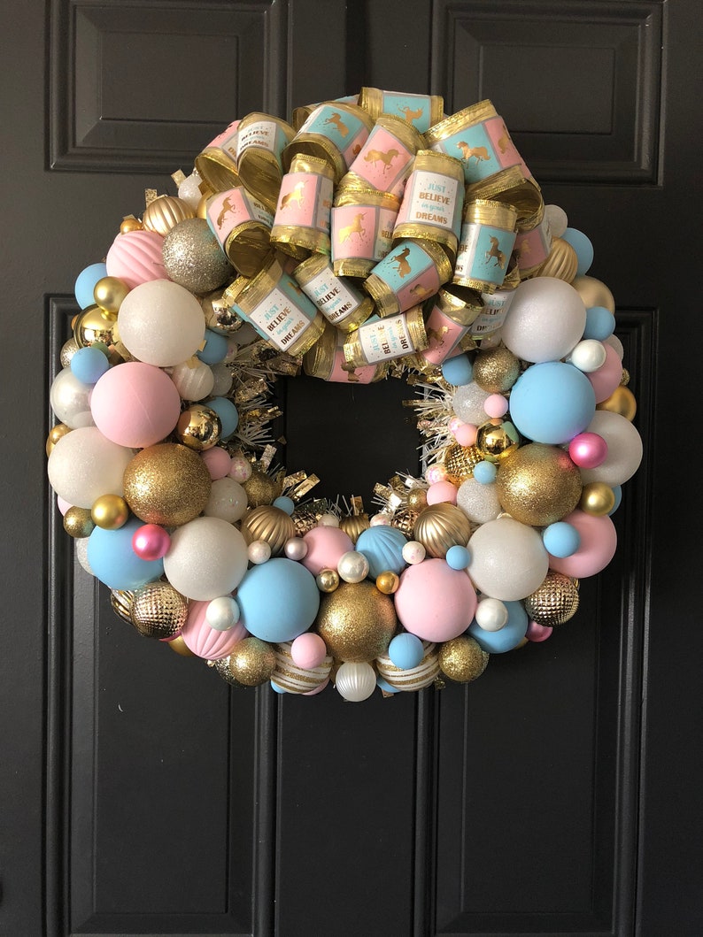 May include: A pink and blue Christmas wreath decorated with gold ornaments and a large bow with the text "Just Believe in Your Dreams".