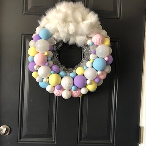 May include: A pastel-colored Christmas wreath with white faux fur on top. The wreath is made of silver tinsel and is decorated with pastel blue, pink, yellow, purple, and white ornaments. The wreath is hanging on a black door with the word "Welcome" written on it.