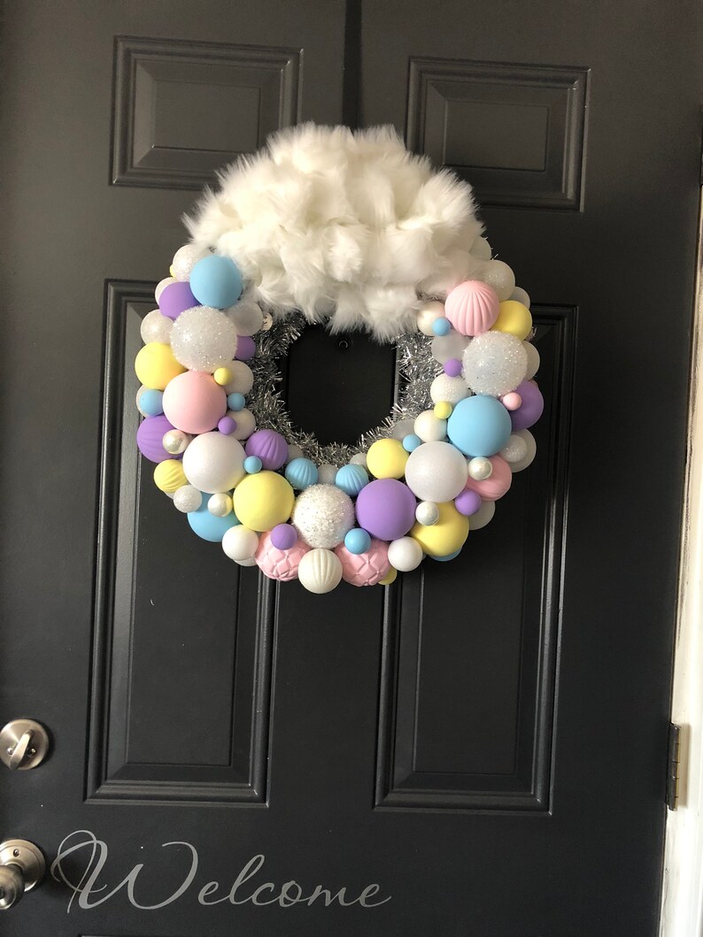 May include: A pastel-colored Christmas wreath with white faux fur on top. The wreath is made of silver tinsel and is decorated with pastel blue, pink, yellow, purple, and white ornaments. The wreath is hanging on a black door with the word "Welcome" written on it.