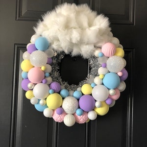 May include: A pastel-colored Christmas wreath with a white faux fur bow. The wreath is decorated with pastel pink, yellow, blue, and purple ornaments. The ornaments are a mix of matte and glitter finishes.