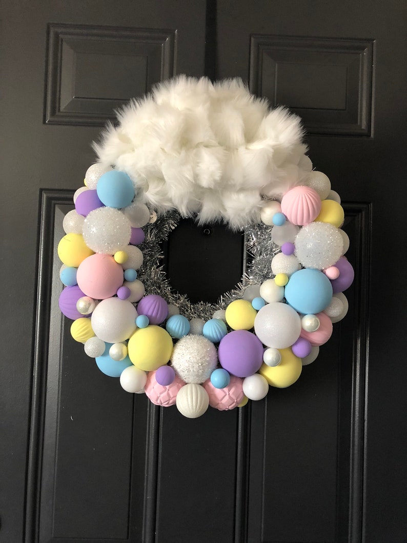May include: A pastel-colored Christmas wreath with a white faux fur bow. The wreath is decorated with pastel pink, yellow, blue, and purple ornaments. The ornaments are a mix of matte and glitter finishes.