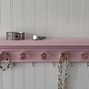 Peut inclure: Une étagère en bois rose avec cinq crochets et des boutons en cristal. L'étagère est ornée d'une garniture décorative et est décorée de bijoux, dont une montre, un bracelet et des colliers.