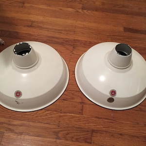 May include: Two white metal industrial pendant light shades with black rims and red labels. The shades are sitting on a wooden floor.