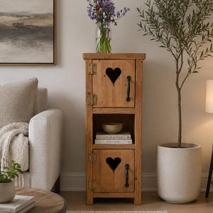 May include: A wooden cabinet with a natural finish and heart-shaped cutouts on the doors. The cabinet has black handles and brass hinges. A vase of purple flowers sits on top. A small olive tree is to the right.
