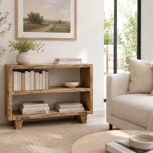 May include: A rectangular wooden bookshelf with a natural light brown finish and visible wood grain. The unit features two horizontal shelves supported by four short, block-style feet. The shelves are filled with various books and a small bowl.