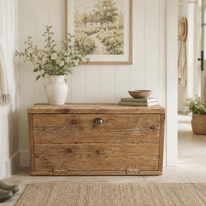 May include: A weathered wooden chest with a distressed finish, holding a white vase of flowers, books, and a wooden bowl. Olive green wellington boots are placed nearby. A framed landscape painting hangs on the wall.