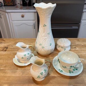 May include: A cream-coloured ceramic vase, two small jugs, a teacup and saucer, and a covered dish, all decorated with green shamrocks. The vase is tall and slender, while the other pieces are smaller and rounder. The set is arranged on a wooden surface.