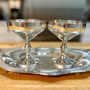 Op de afbeelding: Twee zilveren champagnecoupeglazen op een bijpassend zilveren dienblad. De glazen hebben een klassiek, steelontwerp met een brede, ondiepe kom. De lade heeft een sierlijke, decoratieve rand. De items worden op een houten oppervlak weergegeven.