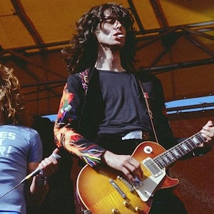 May include: A light blue t-shirt with the text NURSES DO IT BETTER in white letters, worn by a person with long curly blonde hair, standing next to a person with long dark hair playing a sunburst electric guitar with a colorful patterned sleeve.