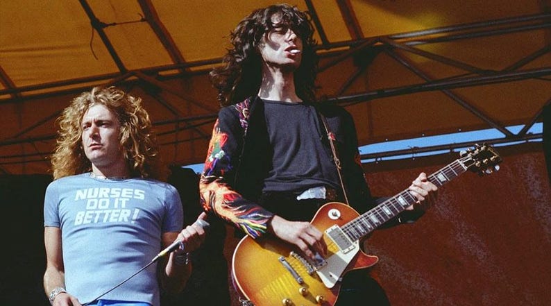 May include: A light blue t-shirt with the text NURSES DO IT BETTER in white letters, worn by a person with long curly blonde hair, standing next to a person with long dark hair playing a sunburst electric guitar with a colorful patterned sleeve.