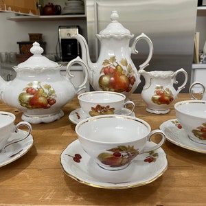 Puede incluir: Un juego de té de porcelana blanca con un motivo de frutas. El juego incluye una tetera, una jarra de crema, un azucarero y cuatro tazas de té y platillos. El motivo de la fruta presenta peras, manzanas y uvas en tonos de rojo, verde y morado.
