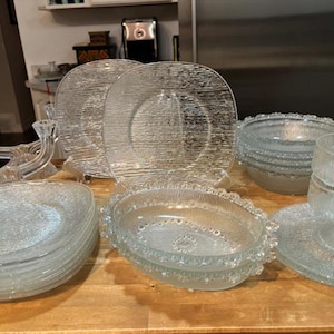 May include: A set of clear glass plates, bowls, and serving dishes. The plates have a textured surface and the bowls have a beaded edge.